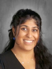 A woman with long hair wearing a black shirt, standing confidently with a neutral background.