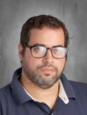 A man wearing glasses and a blue shirt, looking thoughtfully into the distance.