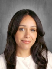 A woman with long hair wearing a white shirt, standing confidently against a neutral background.