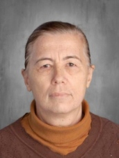 An older woman wearing a brown sweater and a brown scarf, smiling gently.