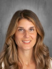 A woman with long hair wearing a white shirt, standing confidently with a serene expression.