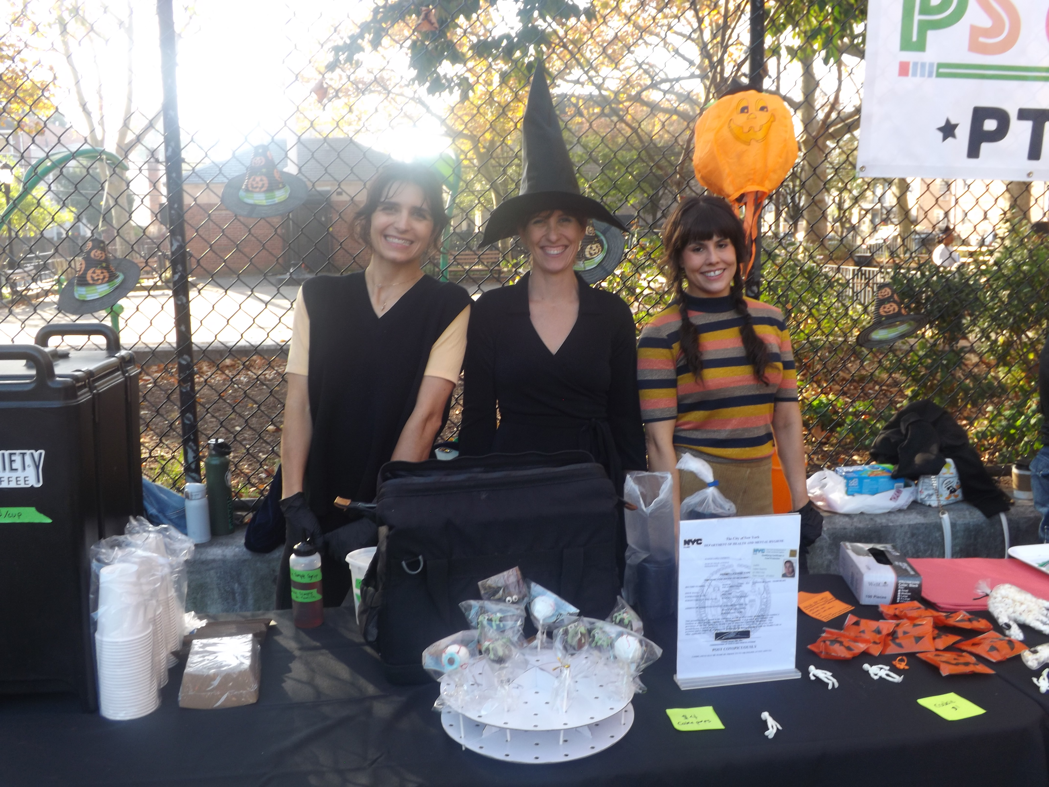 Three women in costumes stand behind a table with a pumpkins and displays.